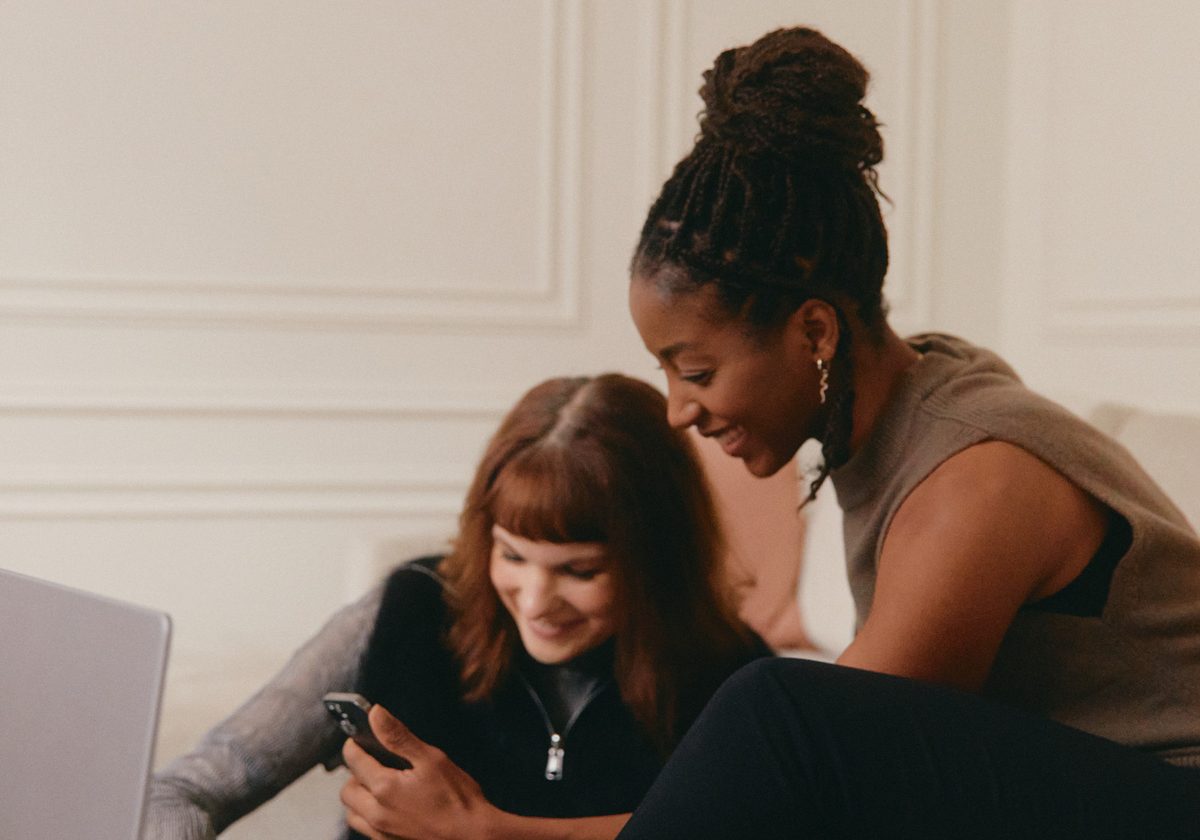 Two women on a couch looking at a phone beside a laptop