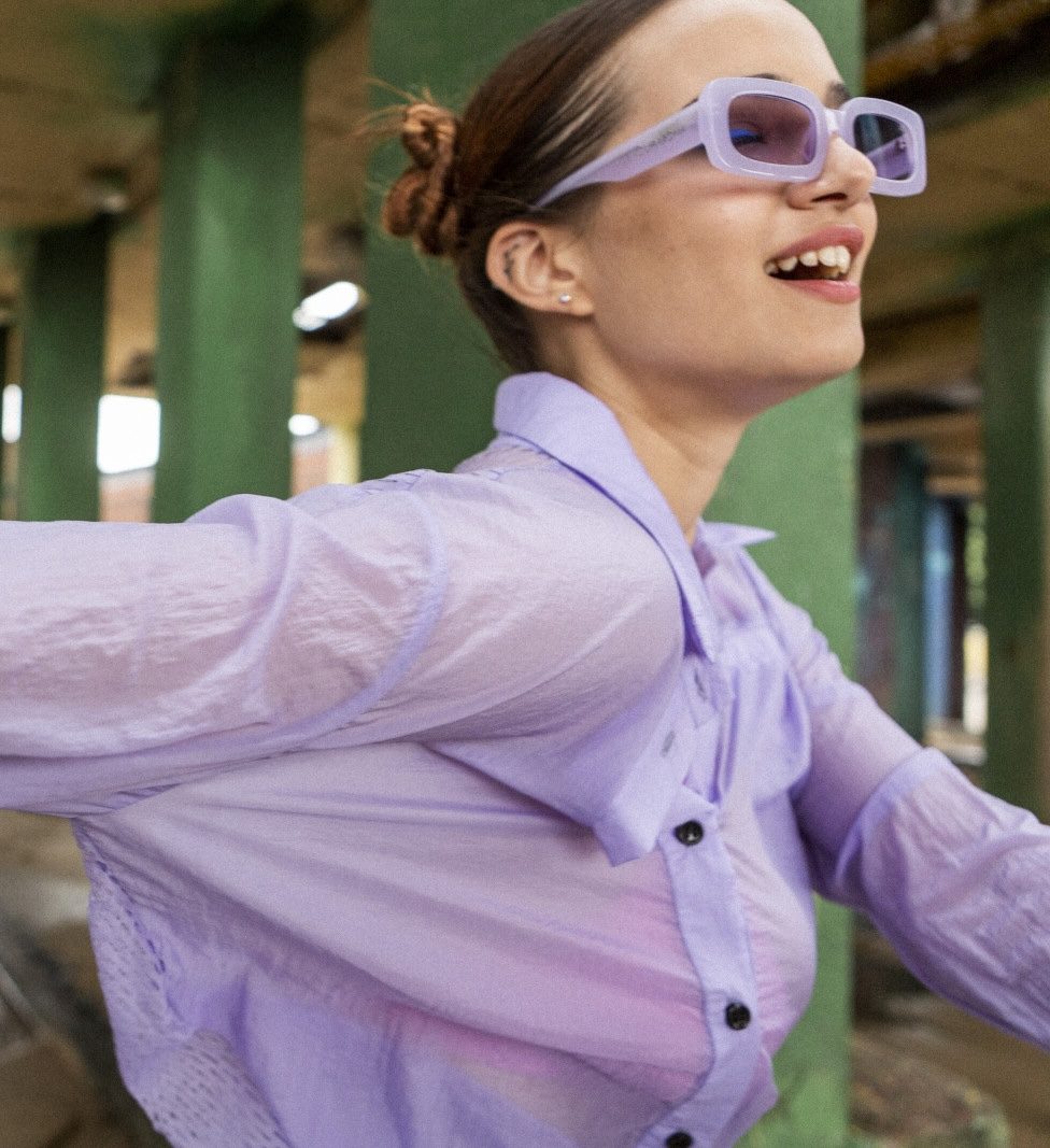 A woman in a lilac shirt and sunglasses, laughing, arms out
